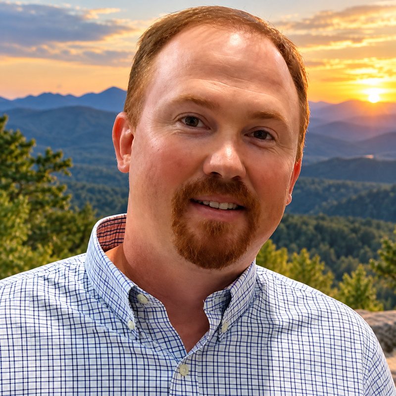 John Morrison, smiling, in a blue checked button-down shirt, with forested mountain ridges and a sunset behind him.